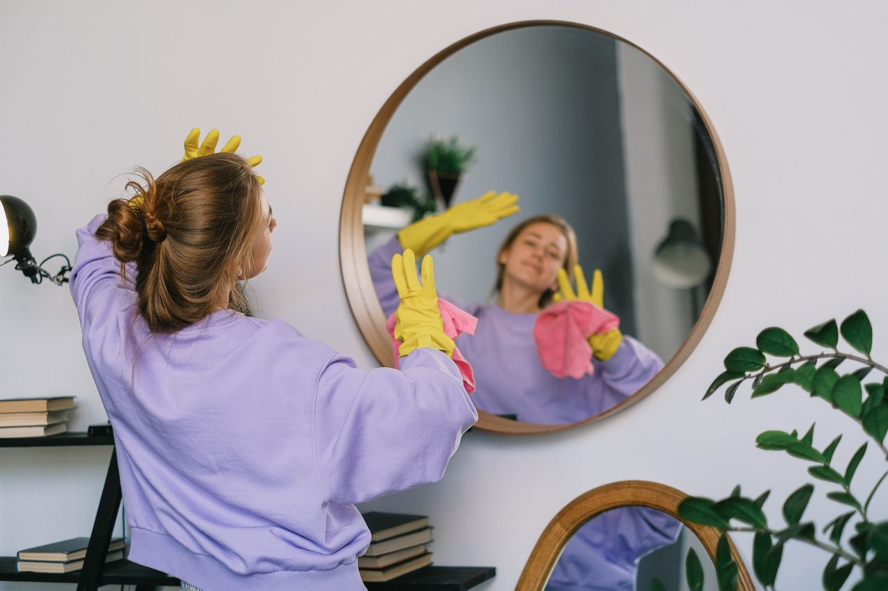 Back view of young female in casual clothes and latex gloves standing against mirror and dancing while cleaning light room in daytime